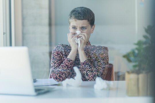 Ill businesswoman at desk in office blowing nose