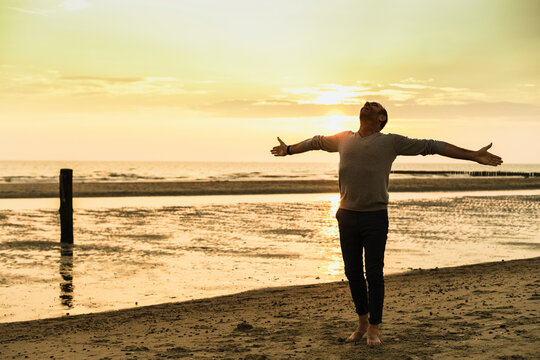Carefree man with arms outstretched standing at beach against sky during sunset