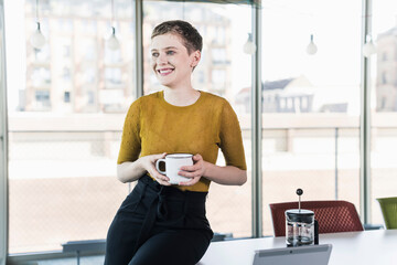 Smiling businesswoman sitting on desk in office holding coffee mug