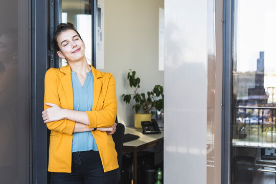 Young businesswoman with arms crossed and eyes closed standing by door at office