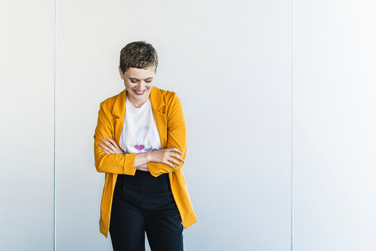 Smiling businesswoman wearing yellow blazer with arms crossed standing against wall in office