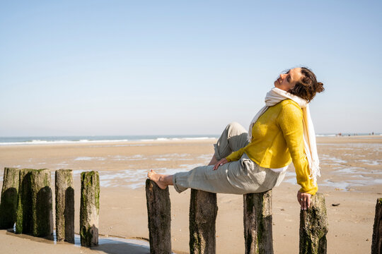 Mature woman with eyes closed sitting on wooden post at beach against clear sky