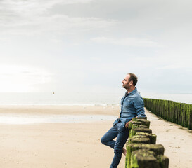 Thoughtful man standing by wooden posts at beach against sky during sunset