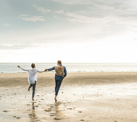 Cheerful mature couple holding hands while jumping at beach against cloudy sky