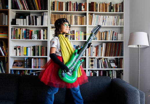 Happy boy dressed up as a rock star standing on the couch at home with inflatable toy guitar