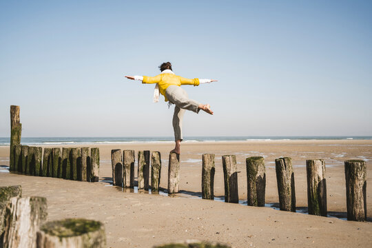 Woman with arms outstretched balancing on wooden posts at beach against clear sky