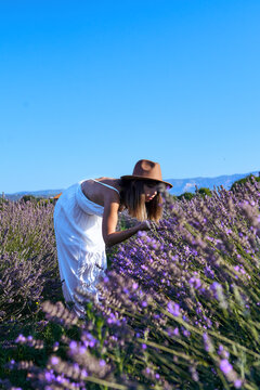 Woman wearing white dress smelling lavender flower while standing in field against clear sky