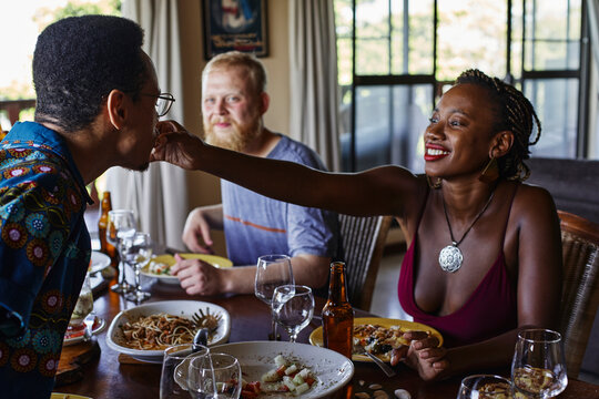 Smiling Young Woman Feeding Friend While Sitting At Dining Table