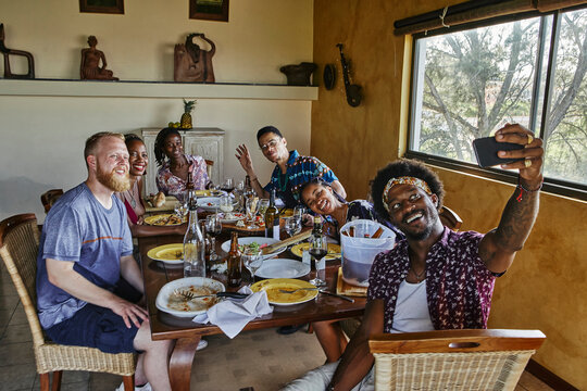 Smiling man taking selfie with male and female friends sitting at dining table during lunch