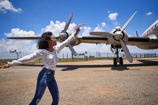 Cheerful young woman holding paper plane against propellers at airport on sunny day