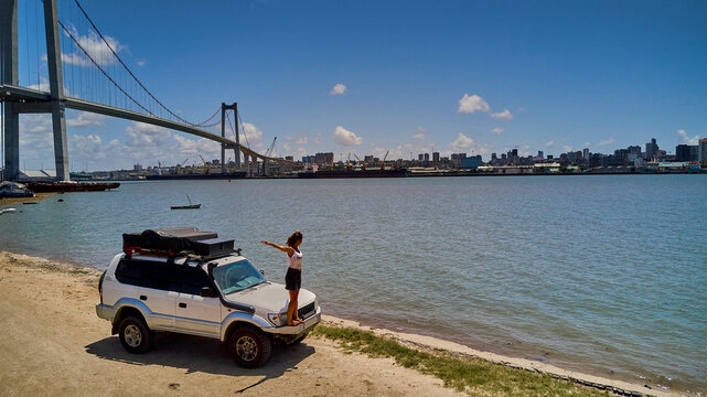 Mozambique, Katembe, Adult woman standing with raised arms on bumper of 4x4 car admiring view of Maputo Bay with city and Maputo-Katembe Bridge in background
