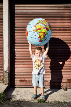 Boy carrying globe while standing on street against closed shutter during sunny day