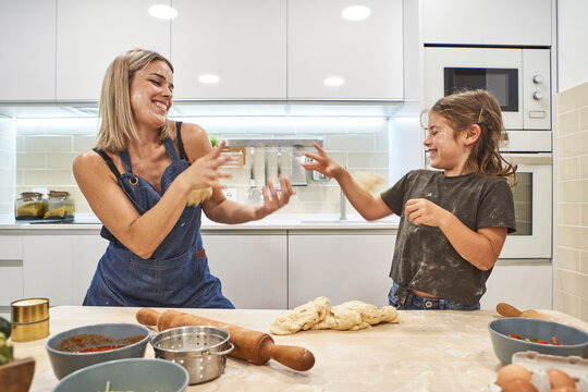 Cheerful mother and daughter playing with pizza dough in kitchen