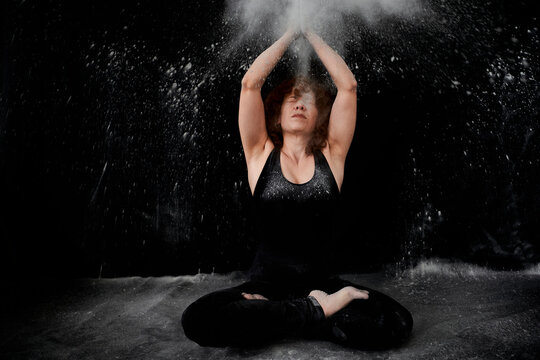 Woman sitting on floor while clapping with white dust over herself against black background