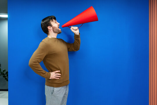 Businessman with hand on hip screaming in megaphone by blue wall at work place