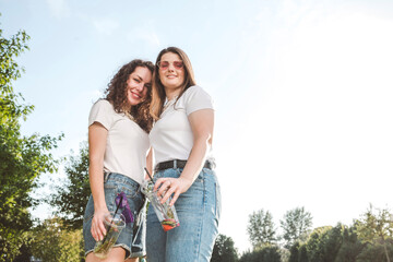 Smiling beautiful women standing with disposable cups at park against sky on sunny day
