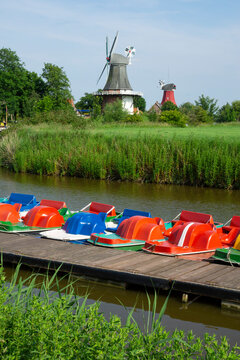 Germany, Lower Saxony, Krummhorn, Pedal boats moored along riverbank jetty with windmills in background