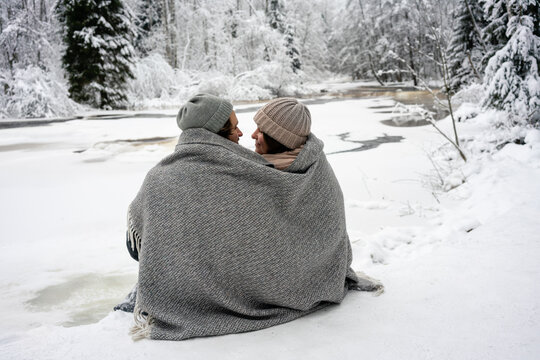 Young couple covered in blanket looking at each other while sitting by frozen river in forest