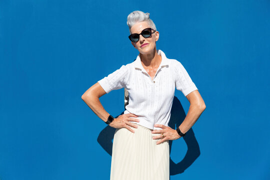 Senior woman standing with hand on hip against blue wall during sunny day