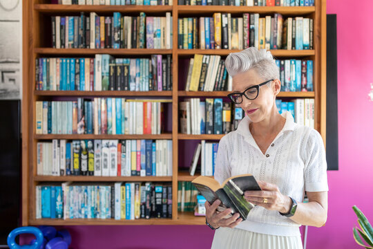 Active senior woman reading book while standing against bookshelf at home
