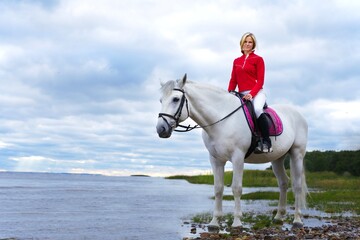 Portrait of a beautiful girl, young woman rider, equestrian on white horse in polo shirt, riding outdoors near lake or beach gulf. Horseback riding sport, natural background. 