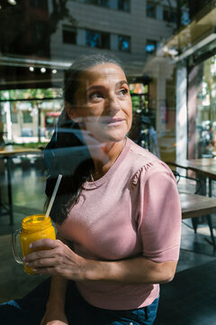Thoughtful woman holding mason jar with juice while looking through window at cafe