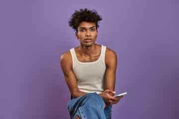 Close-up portrait of young man sits with mobile and looking camera on isolated purple background