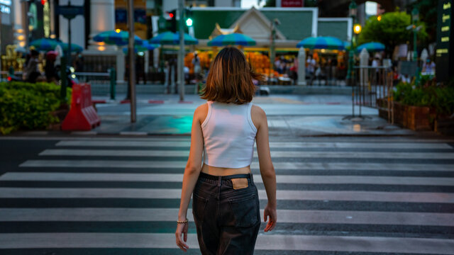 Young beautiful Asian woman walking on street crosswalk in the city and looking at crowd of people and illuminated night lights. Pretty girl enjoy urban outdoor activity lifestyle and city nightlife.