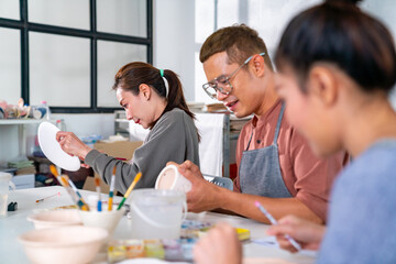 Group of Asian man and woman millennial generation friends enjoy and having fun painting self-made pottery together at pottery studio. Hobbies and leisure activity handmade ceramic painting workshop. - obrazy, fototapety, plakaty