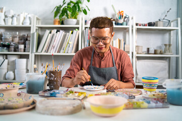 Fototapeta na wymiar Asian LGBTQ guy learning color painting her self-made pottery at home. Confidence male enjoy hobbies and indoors leisure activity handicraft ceramic sculpture and painting workshop at pottery studio