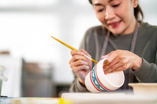 Asian woman learning color painting her self-made pottery at home. Confidence female enjoy hobbies and indoors leisure activity handicraft ceramic sculpture and painting workshop at pottery studio.
