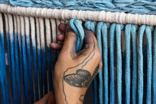 Mano tatuada de mujer trabajando pieza de macram&eacute; te&ntilde;ido de azul.