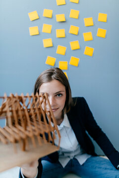 Young businesswoman holding architectural model with yellow sticky notes on the wall behind ger