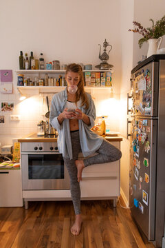 Young woman in yoga pose checking cell phone in kitchen at home