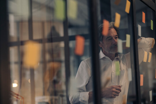 Senior businessman looking at adhesive notes at glass pane in office