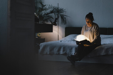 Senior woman sitting on bed holding glowing book