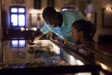 Afro male visitor and his little daughter visiting museum and looking at showcase with exhibits