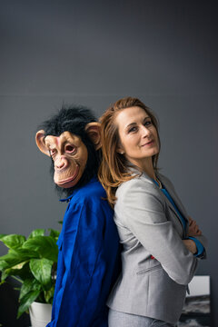Businesswoman standing fback to back with a trainee, wearing monkey mask and blue oevrall