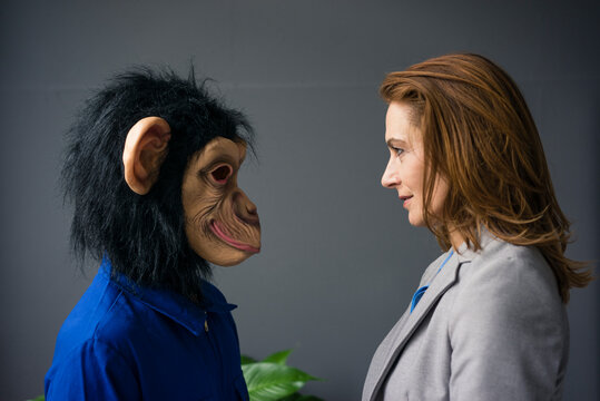 Businesswoman standing face to face with a trainee, wearing monkey mask and blue oevrall