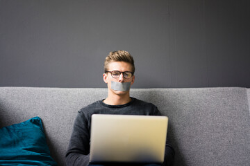 Young man with taped mouth sitting on couch using laptop