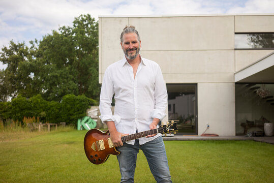Portrait of mature man standing on lawn in front of his house holding guitar