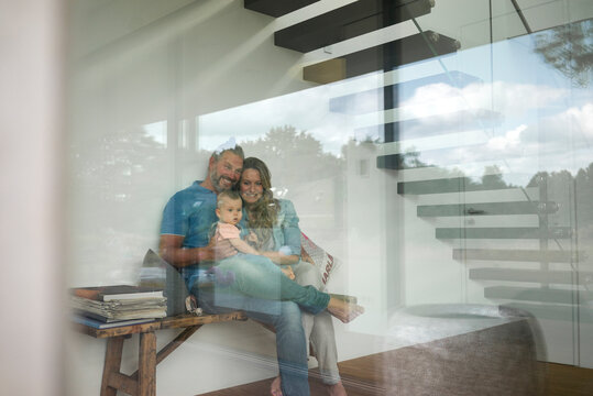 Happy family of three sitting on a bench behind windowpane at home