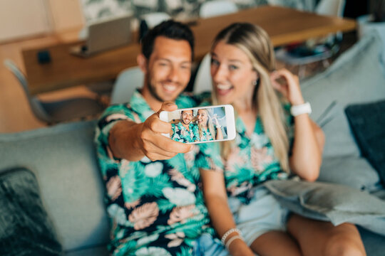 Happy couple sitting on couch in living room wearing Hawaiian shirts taking a selfie
