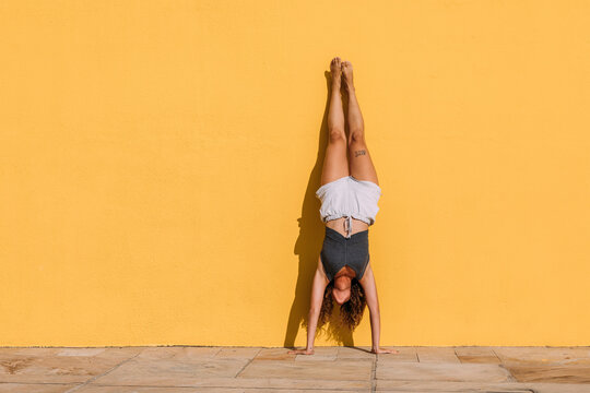 Young woman doing a handstand in front of a yellow wall