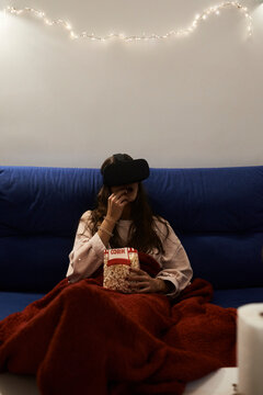 Young woman using VR headsets eating popcorn on the sofa at home at night