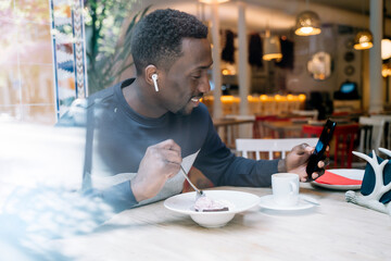 Smiling young man with earphones and smartphone behind windowpane in a restaurant