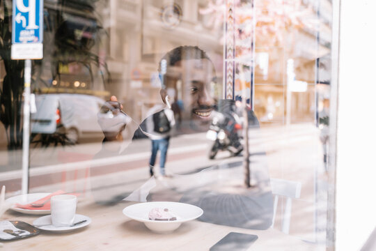 Smiling young man with wireless earphones behind windowpane in a coffee shop eating pastry