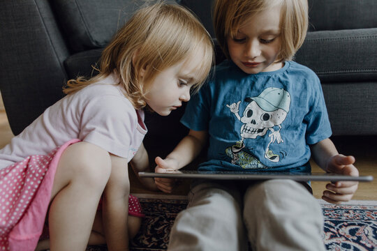 Brother and little sister sitting on the floor at home using digital tablet
