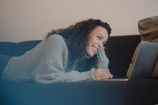 Woman lying on couch, using laptop, smiling