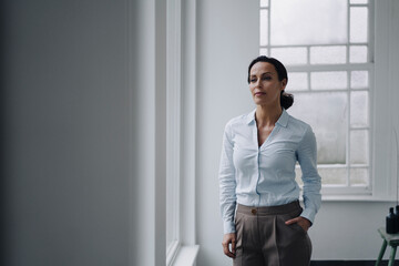 Successful businesswoman, standing by window, with hand in pocket